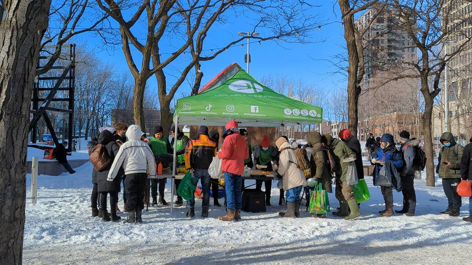 Volunteers in green vests serve under the tent in the snow while people in winter coats wait their turn, seen from behind.