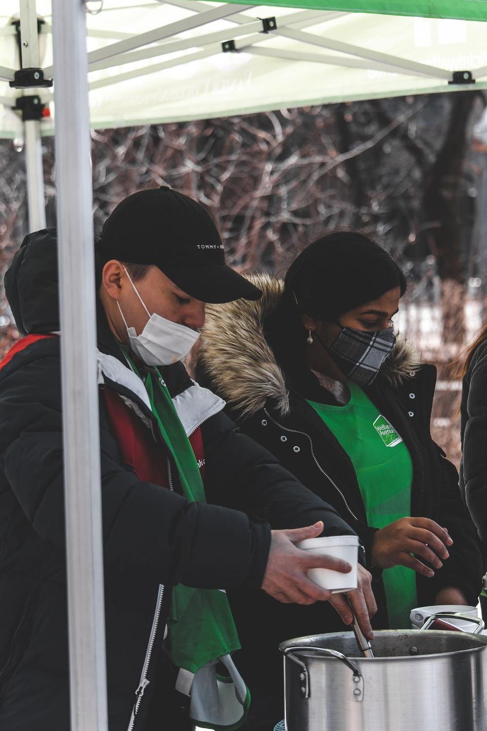 Two masked volunteers in green vests ladle soup from a large pot into cups under the tent.