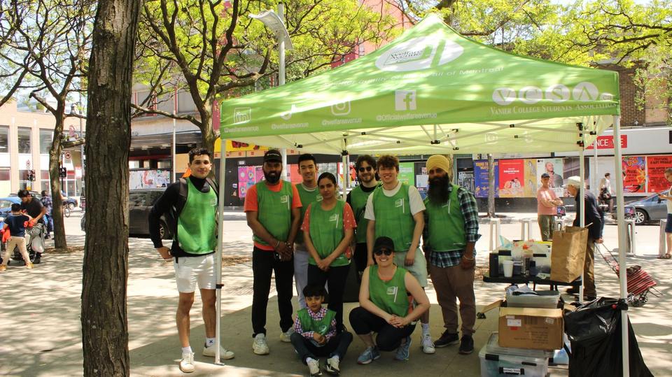 About twelve volunteers, most in green vests, pose in front of the green tent in a downtown park.