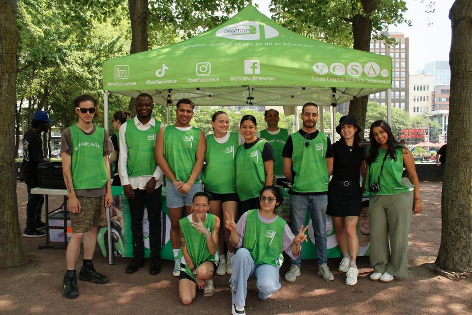 Thirteen volunteers in green Welfare Avenue vests pose under the green tent in a leafy park.